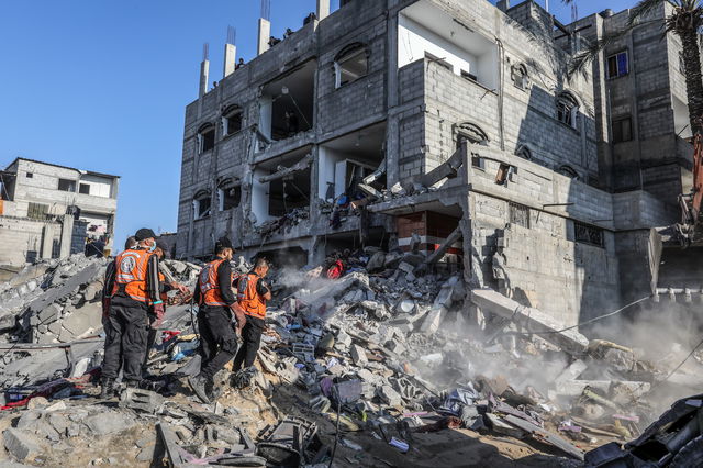 Ein zerstörtes Wohnhaus, Trümmer, dahinter Reste des Hauses, davor Rettungskräfte in orangen Warnwesten