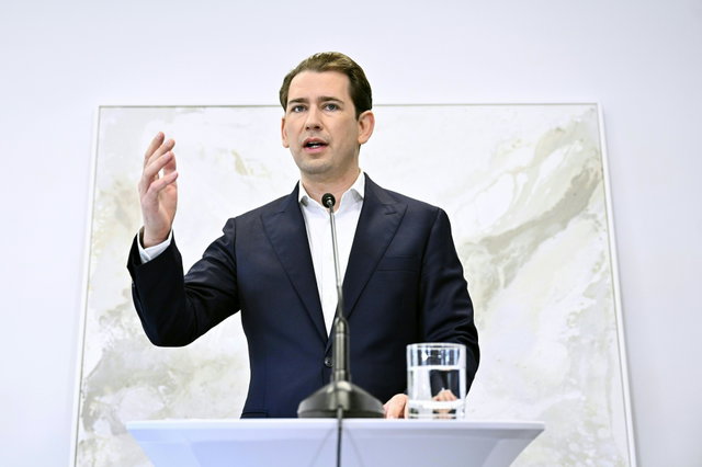 (Dieser ALT-Text wurde mithilfe von KI erstellt) Mann im Anzug spricht an einem Rednerpult mit Mikrofon, vor hellem Hintergrund, ein Glas Wasser steht auf dem Pult.