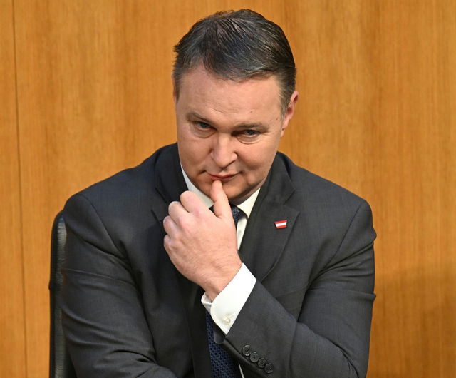 (Dieser ALT-Text wurde mithilfe von KI erstellt) Mann im dunklen Anzug sitzt vor einer Holzvertäfelung und blickt nachdenklich, mit der Hand am Kinn.