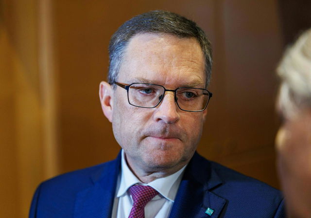 (Dieser ALT-Text wurde mithilfe von KI erstellt) Mann mit Brille und Anzug blickt ernst nach unten, unscharfer Hintergrund, Gesprächspartner am rechten Bildrand angeschnitten.