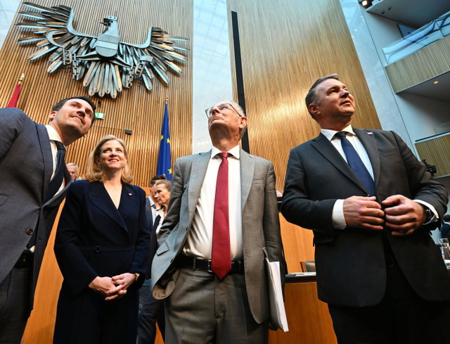 (Dieser ALT-Text wurde mithilfe von KI erstellt) Vier Personen in Anzügen stehen in einem modernen Sitzungssaal unter dem österreichischen Bundesadler und vor einer EU-Flagge.