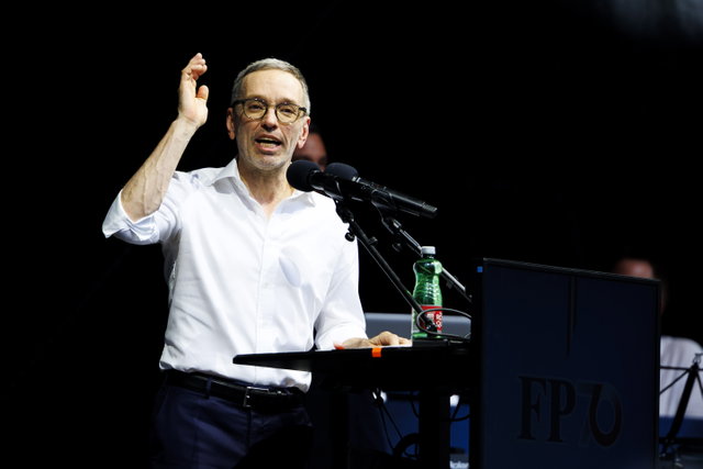 (Dieser ALT-Text wurde mithilfe von KI erstellt) Mann mit Brille spricht an einem Rednerpult mit Mikrofonen und Wasserflasche, auf dem Pult steht „FPÖ“.