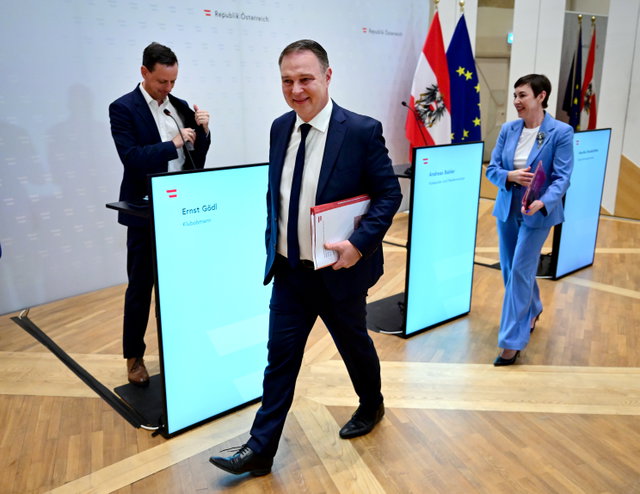 (Dieser ALT-Text wurde mithilfe von KI erstellt) Drei Personen in Businesskleidung gehen und stehen vor Rednerpulten mit Namensschildern, im Hintergrund österreichische und EU-Flaggen.