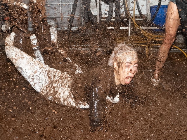 (Dieser ALT-Text wurde mithilfe von KI erstellt) Person in weißer Kleidung fällt in eine große Menge Schlamm, während Schlamm spritzt; industrielle Umgebung im Hintergrund.
