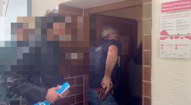 (Dieser ALT-Text wurde mithilfe von KI erstellt) Ein Polizist mit sichtbarer Waffe betritt ein Gebäude, weitere Personen im Bild sind verpixelt und nicht erkennbar.