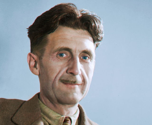 (Dieser ALT-Text wurde mithilfe von KI erstellt) Porträt eines Mannes mit braunen Haaren, Schnurrbart und braunem Jackett vor einfarbig blauem Hintergrund.