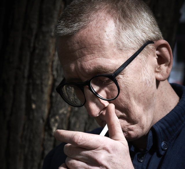 (Dieser ALT-Text wurde mithilfe von KI erstellt) Nahaufnahme eines älteren Mannes mit Brille, der im Freien eine Zigarette anzündet. Im Hintergrund ist ein Baumstamm zu sehen.