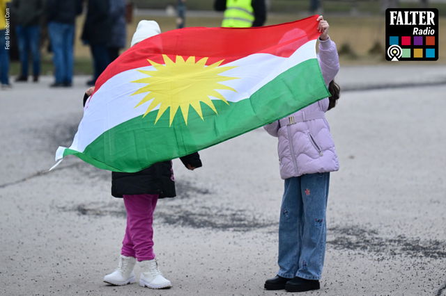 (Dieser ALT-Text wurde mithilfe von KI erstellt) Zwei Kinder halten gemeinsam eine kurdische Flagge mit rotem, weißem und grünem Streifen sowie gelber Sonne in der Mitte.
