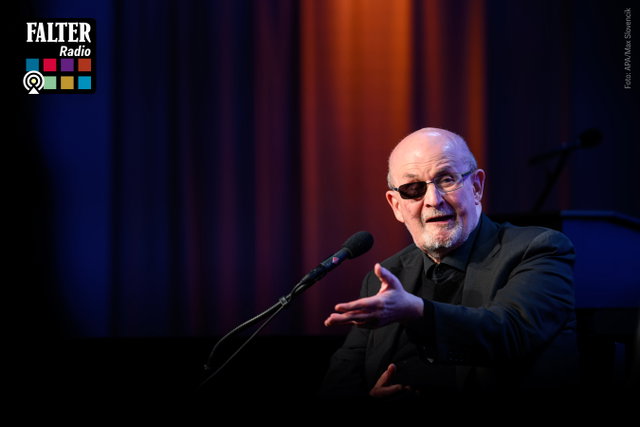 (Dieser ALT-Text wurde mithilfe von KI erstellt) Älterer Mann mit Brille spricht auf einer Bühne in ein Mikrofon; oben links Logo „FALTER Radio“ mit bunten Quadraten.