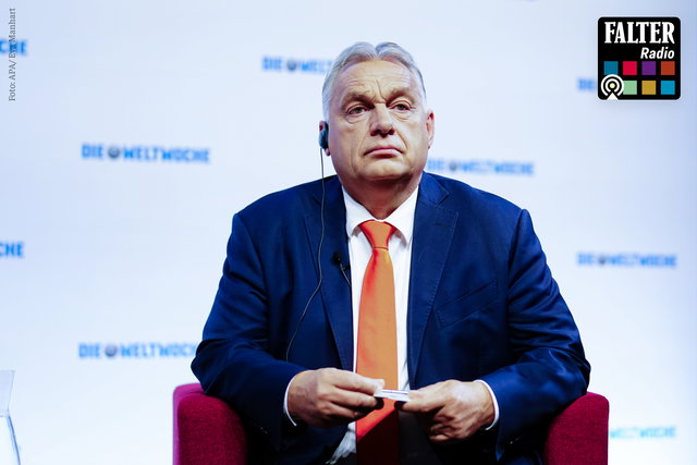 (Dieser ALT-Text wurde mithilfe von KI erstellt) Mann im blauen Anzug mit roter Krawatte sitzt auf einem roten Stuhl vor Hintergrund mit „DIE WELTWOCHE“-Logo; oben rechts „FALTER Radio“-Logo.