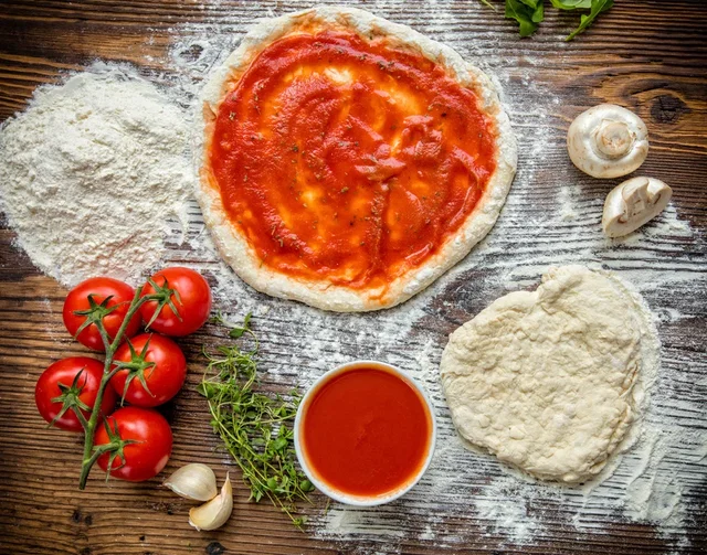 (Dieser ALT-Text wurde mithilfe von KI erstellt) Zutaten für Pizza auf Holz: ausgerollter Teig mit Tomatensauce, roher Teig, Mehl, Tomaten, Kräuter, Knoblauch, Pilze und Schale mit Tomatensauce.