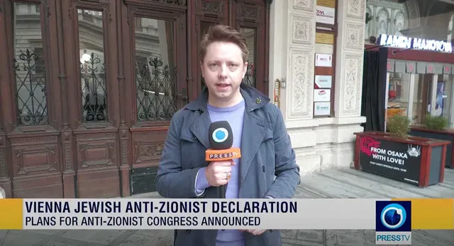 (Dieser ALT-Text wurde mithilfe von KI erstellt) Reporter steht mit PressTV-Mikrofon vor einem Gebäude; eingeblendeter Text: „Vienna Jewish Anti-Zionist Declaration – Plans for Anti-Zionist Congress Announced“.