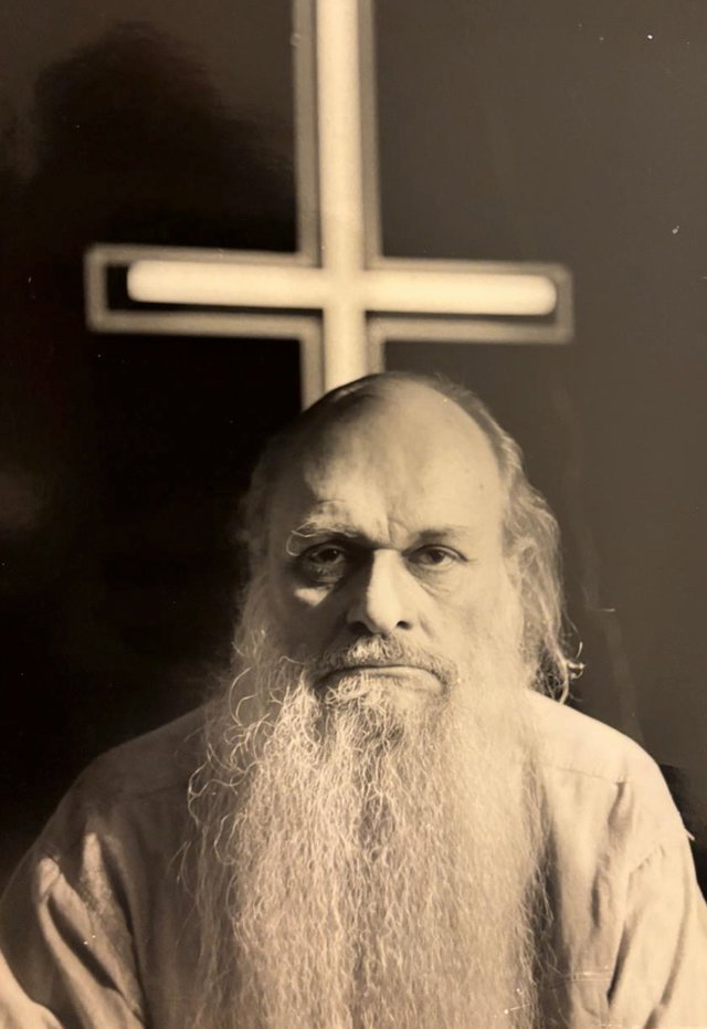(Dieser ALT-Text wurde mithilfe von KI erstellt) Schwarz-weiß-Fotografie eines älteren Mannes mit langem Bart vor einem beleuchteten Kreuz an der Wand.