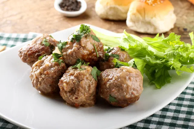 (Dieser ALT-Text wurde mithilfe von KI erstellt) Sechs gebratene Fleischbällchen mit Petersilie auf einem weißen Teller, daneben frischer grüner Salat und im Hintergrund Brötchen.