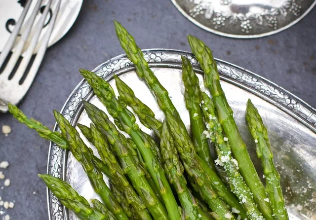 (Dieser ALT-Text wurde mithilfe von KI erstellt) Grüner Spargel mit grobem Salz auf einem silbernen Teller, daneben Besteck auf dunkler Oberfläche.