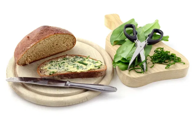 (Dieser ALT-Text wurde mithilfe von KI erstellt) Ein Brotlaib und eine Scheibe Brot mit Kräuterbutter auf einem Holzbrett, daneben Bärlauch, Kräuterschere und Messer auf einem zweiten Brett.