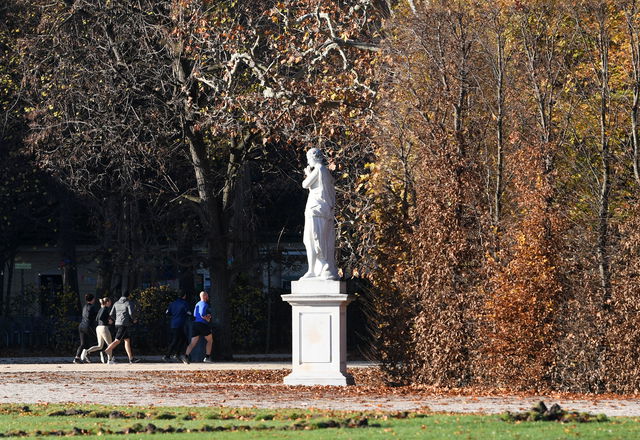 (Dieser ALT-Text wurde mithilfe von KI erstellt) Weiße Statue auf Sockel im herbstlichen Park, im Hintergrund joggen mehrere Personen auf einem Weg entlang.