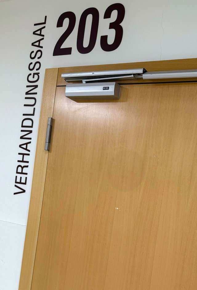 (Dieser ALT-Text wurde mithilfe von KI erstellt) Holztür mit Türschließer, darüber die Aufschrift „203“ und seitlich „Verhandlungssaal“ an der Wand.