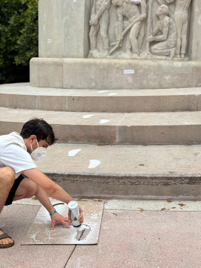 (Dieser ALT-Text wurde mithilfe von KI erstellt) Person mit Maske sprüht mithilfe einer Schablone weiße Fußabdrücke auf den Boden vor einem steinernen Denkmal mit Relief.