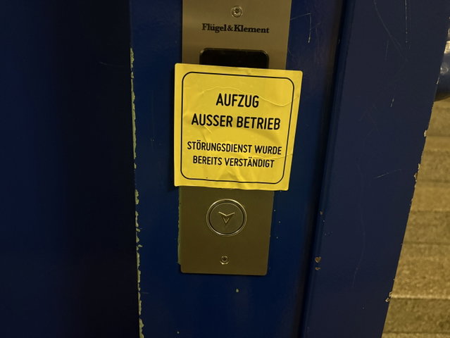 (Dieser ALT-Text wurde mithilfe von KI erstellt) Gelbes Schild an einem Aufzug mit der Aufschrift: „Aufzug außer Betrieb. Störungsdienst wurde bereits verständigt.“