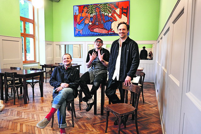 (Dieser ALT-Text wurde mithilfe von KI erstellt) Drei Männer sitzen und stehen in einem hellen Café mit Holzboden, Holztischen und einem bunten Gemälde an der Wand.