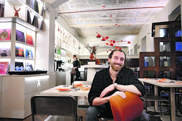(Dieser ALT-Text wurde mithilfe von KI erstellt) Ein Mann sitzt lächelnd an einem Tisch in einem modern eingerichteten Restaurant mit Plattenregalen und dekorativen roten Lampen.