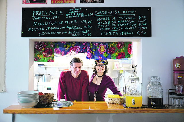 (Dieser ALT-Text wurde mithilfe von KI erstellt) Zwei Personen stehen lächelnd hinter einer Theke in einem Restaurant; darüber eine Tafel mit Speisekarte auf Portugiesisch und Deutsch.
