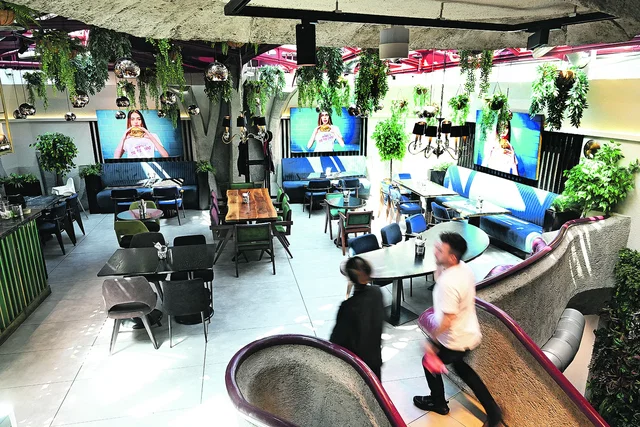 (Dieser ALT-Text wurde mithilfe von KI erstellt) Modernes Restaurant mit vielen Pflanzen, bunten Stühlen, großen Bildschirmen an den Wänden und zwei Personen, die eine Rutsche im Vordergrund benutzen.