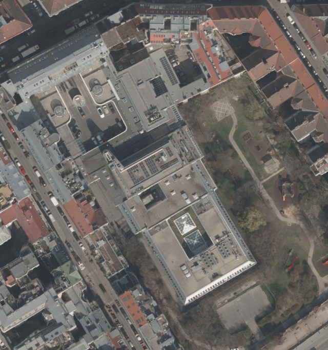 (Dieser ALT-Text wurde mithilfe von KI erstellt) Luftbild zeigt ein städtisches Gebäude mit Flachdach, umgeben von Straßen, Wohnhäusern und einer angrenzenden Parkanlage mit Wegen und Bäumen.