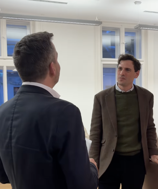 (Dieser ALT-Text wurde mithilfe von KI erstellt) Zwei Männer im Gespräch in einem hellen Büroraum mit weißen Wänden und großen Fenstern im Hintergrund.