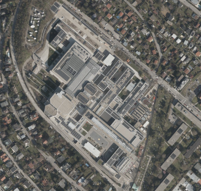 (Dieser ALT-Text wurde mithilfe von KI erstellt) Luftaufnahme eines großen Gebäudekomplexes mit umliegenden Straßen und Wohnhäusern in einem städtischen Gebiet.