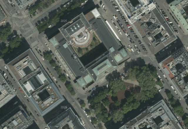 (Dieser ALT-Text wurde mithilfe von KI erstellt) Satellitenbild einer städtischen Umgebung mit mehreren Gebäuden, Straßen, parkenden Autos und einer größeren Grünfläche mit Bäumen.