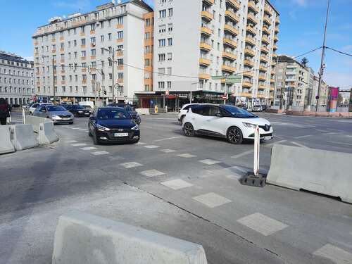 (Dieser ALT-Text wurde mithilfe von KI erstellt) Mehrspurige Straßenkreuzung mit Autos, Betonabsperrungen und Zebrastreifen vor Wohngebäuden in einer Stadt.