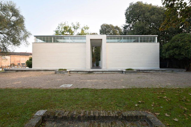(Dieser ALT-Text wurde mithilfe von KI erstellt) Frontansicht eines modernen, weißen Pavillons mit rechteckiger Form, großen Glasfenstern oben und zentralem Durchgang, umgeben von Bäumen und Kiesfläche.