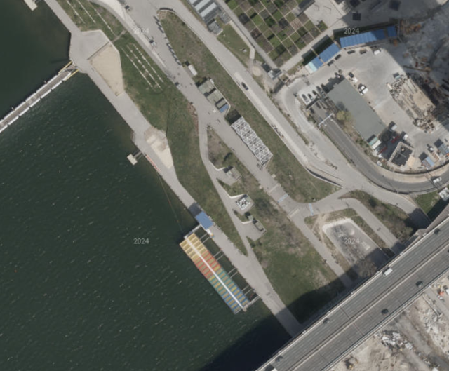 (Dieser ALT-Text wurde mithilfe von KI erstellt) Satellitenbild eines Uferbereichs mit Straßen, Grünflächen, Gebäuden und einer bunten Steganlage am Wasser.