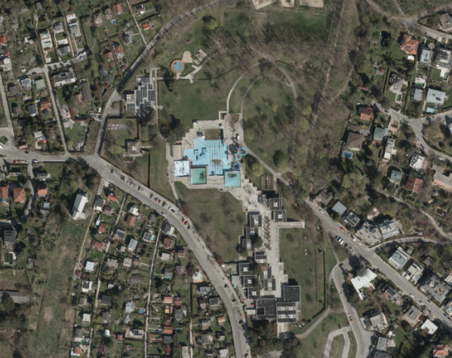 (Dieser ALT-Text wurde mithilfe von KI erstellt) Luftbild zeigt ein städtisches Gebiet mit Gebäuden, Straßen, Grünflächen und einem zentralen Gebäudekomplex mit blauen Dächern.