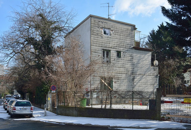 (Dieser ALT-Text wurde mithilfe von KI erstellt) Eckiges Wohnhaus mit grauer Fassade in der Neblingergasse 9, Wien, im Winter; davor schneebedeckter Gehweg und parkende Autos.
