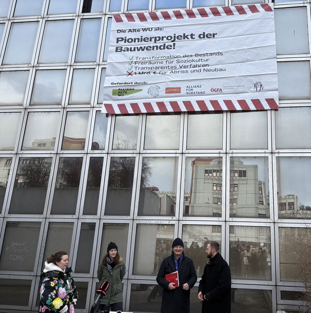 (Dieser ALT-Text wurde mithilfe von KI erstellt) Vier Personen stehen vor einem Gebäude mit großen Fenstern; darüber hängt ein Banner mit dem Text „Pionierprojekt der Bauwende!“ und weiteren Informationen zur Gebäudenutzung.