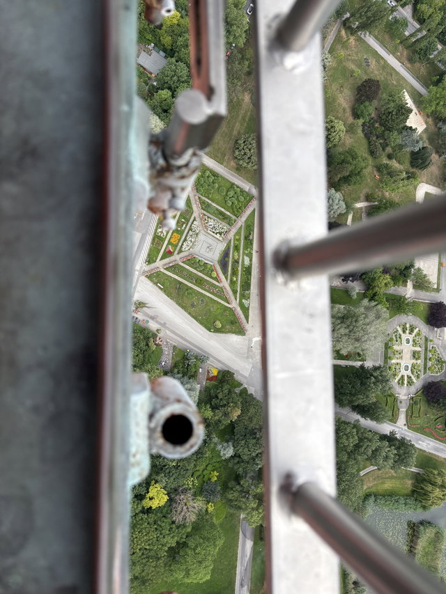 (Dieser ALT-Text wurde mithilfe von KI erstellt) Blick von oben durch ein Metallgeländer auf eine geometrisch angelegte Parkanlage mit Wegen, Blumenbeeten und Grünflächen.