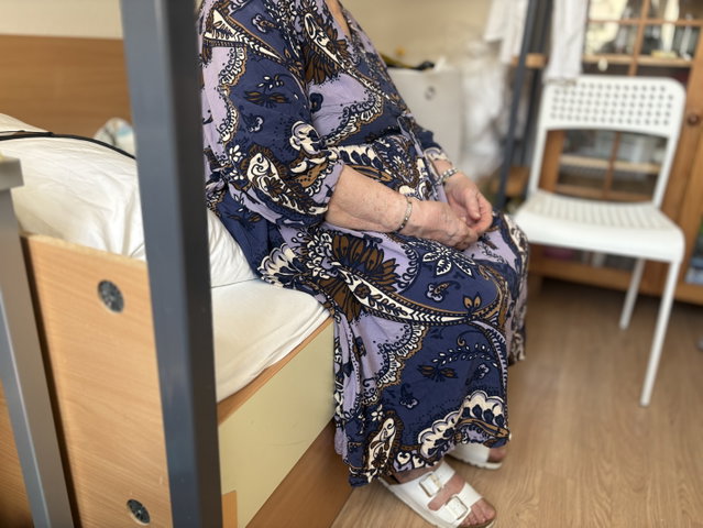 (Dieser ALT-Text wurde mithilfe von KI erstellt) Ältere Person sitzt in gemustertem Kleid auf einem Pflegebett in einem Zimmer mit Holzfußboden und weißem Stuhl im Hintergrund.