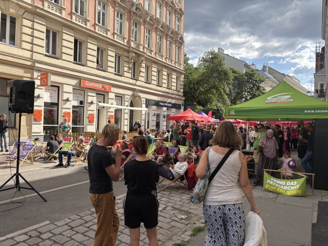 (Dieser ALT-Text wurde mithilfe von KI erstellt) Straßenfest mit vielen Menschen, Sitzgelegenheiten, Infoständen und grünen sowie roten Pavillons vor einem Gebäude mit dem Schild „Gummiboot“.