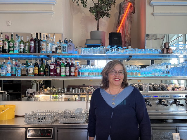 (Dieser ALT-Text wurde mithilfe von KI erstellt) Frau steht lächelnd hinter einer Bar mit Flaschen, Gläsern und Kaffeemaschine; im Hintergrund Statue mit rotem Lichtschwert.