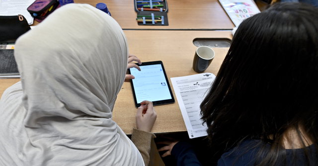 (Dieser ALT-Text wurde mithilfe von KI erstellt) Zwei Schülerinnen sitzen an einem Tisch, arbeiten gemeinsam mit einem Tablet und einem Arbeitsblatt im Klassenraum.