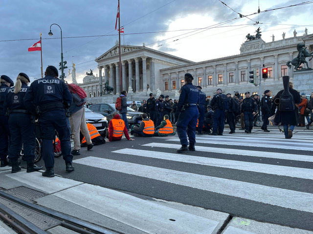 (Dieser ALT-Text wurde mithilfe von KI erstellt) Mehrere Personen mit orangefarbenen Warnwesten sitzen auf einem Zebrastreifen vor dem österreichischen Parlament, umgeben von Polizisten.