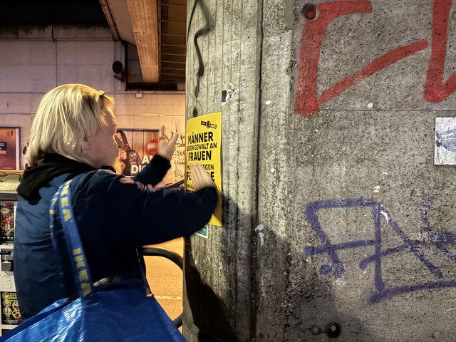 (Dieser ALT-Text wurde mithilfe von KI erstellt) Person klebt ein gelbes Plakat mit der Aufschrift „Männer gegen Gewalt an Frauen“ an eine graue, mit Graffiti besprühte Wand unter einer Brücke.