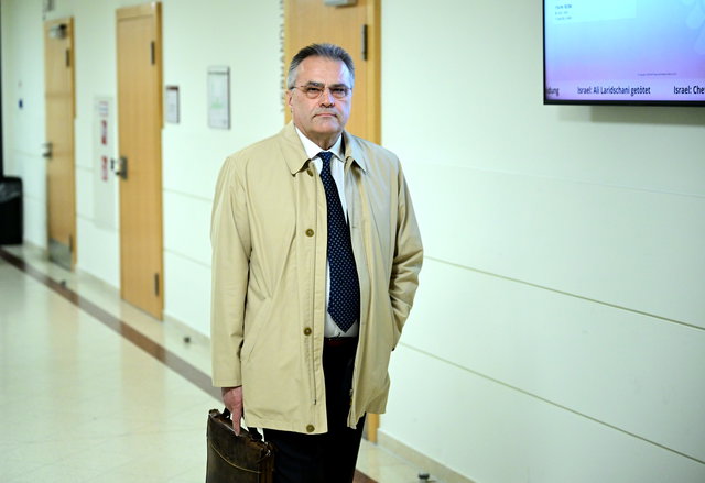 (Dieser ALT-Text wurde mithilfe von KI erstellt) Mann im beigen Mantel mit Aktentasche steht in einem Flur eines Gerichtsgebäudes vor geschlossenen Türen.