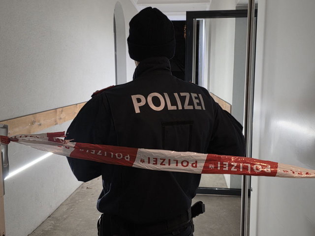 (Dieser ALT-Text wurde mithilfe von KI erstellt) Polizist steht mit dem Rücken zur Kamera hinter einer Absperrung mit „Polizei“-Band in einem Flur.