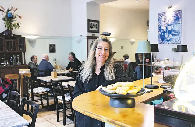 (Dieser ALT-Text wurde mithilfe von KI erstellt) Frau steht lächelnd hinter einer Theke in einem Café, im Vordergrund ein Teller mit Gebäck, im Hintergrund sitzen Gäste an Tischen.