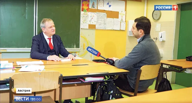 (Dieser ALT-Text wurde mithilfe von KI erstellt) Zwei Männer sitzen sich in einem Klassenzimmer gegenüber, einer hält ein Mikrofon mit der Aufschrift „Россия“, im Hintergrund eine Tafel und Kinderzeichnungen.