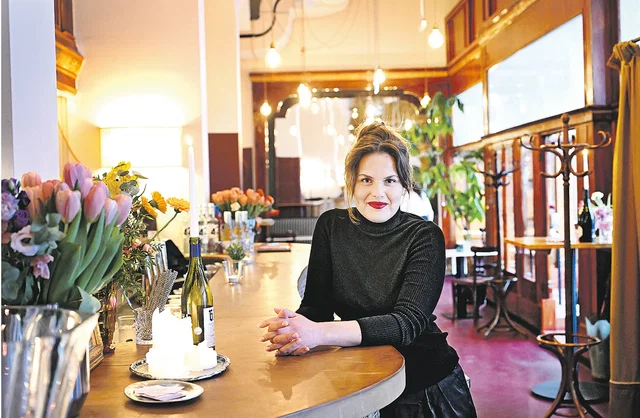 (Dieser ALT-Text wurde mithilfe von KI erstellt) Frau sitzt an einer Theke in einem stilvoll eingerichteten Café mit Blumen, Kerzen und Weinflasche auf dem Tresen.
